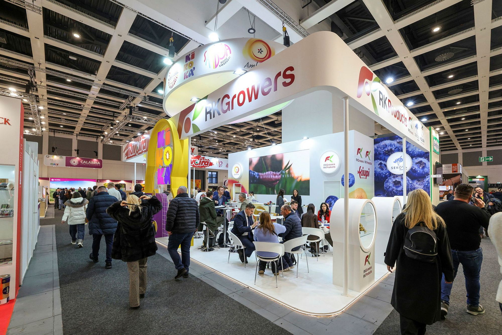 An exhibitor stand with visitors seated at meeting tables on the show floor.