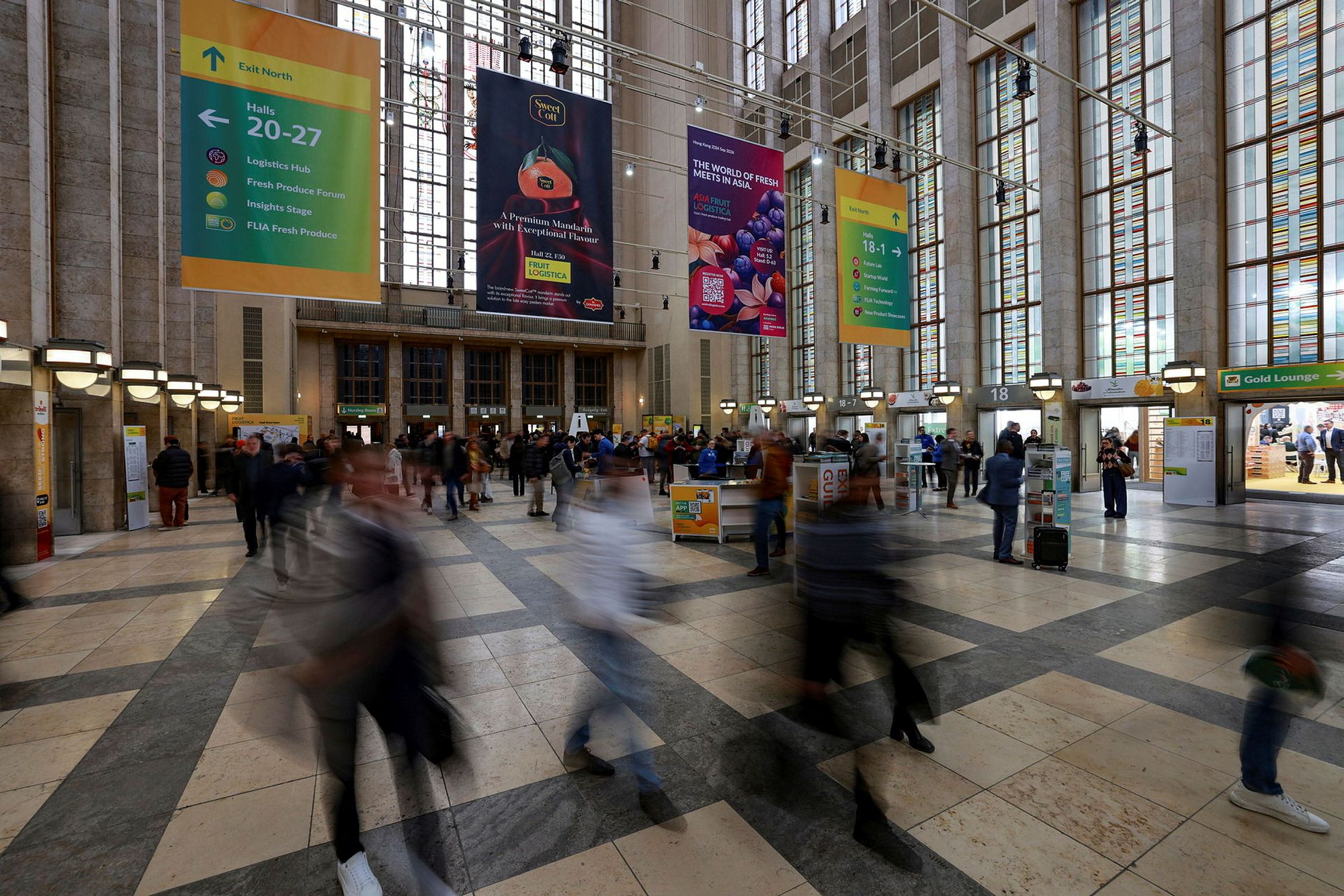 Trade visitors crossing the tall, glass-roofed entrance concourse at FRUIT LOGISTICA in Berlin beneath colourful hall-direction banners.