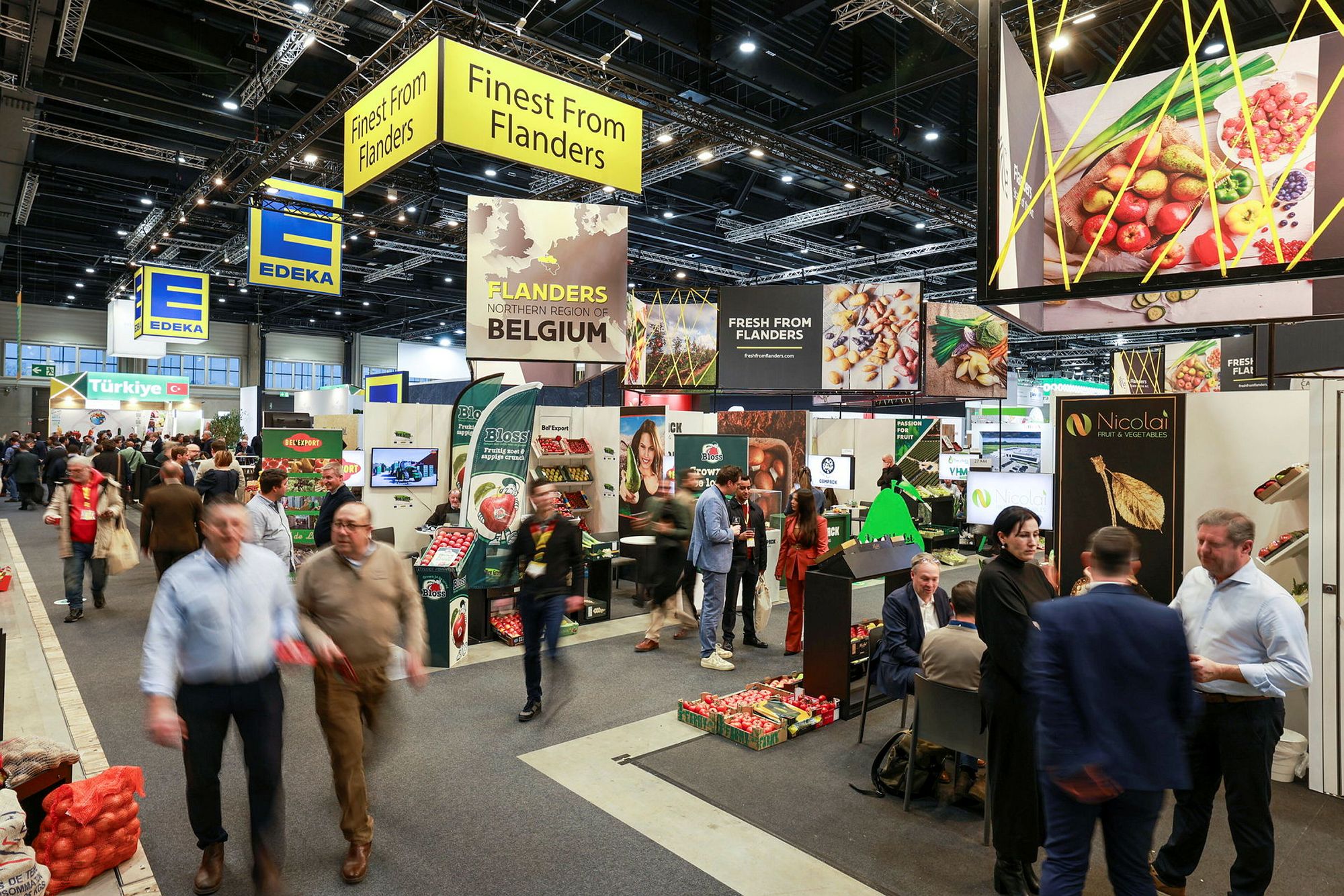 Buyers walking the show floor between country pavilion stands hung with yellow overhead signage.