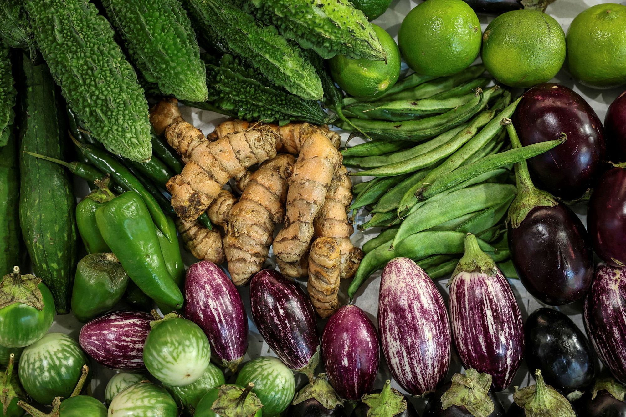 A display of tropical vegetables including turmeric root, striped aubergines, green chillies and limes.